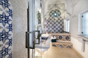 A bathroom with a tiled wall, a sink, a mirror, and a shower area. - Estate4home - Regina Dar (Positano)