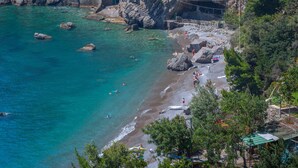 Una playa costera con aguas turquesas cristalinas, terreno rocoso y un edificio de piedra con una escalera que baja a la playa.