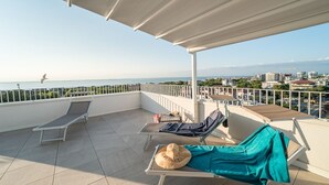A rooftop terrace with two sun loungers, a hat, and a towel, overlooking a cityscape and the sea.
