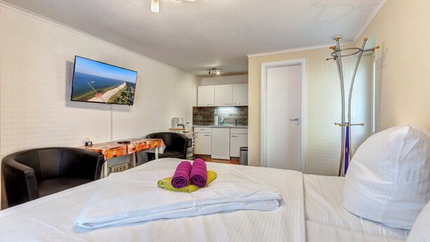 Habitación de hotel con cama, escritorio, silla, televisión y una kitchenette al fondo.