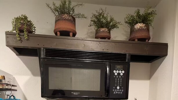 Une cuisine avec un micro-ondes noir, des casseroles suspendues, un sac matelassé bleu et un récipient en métal.