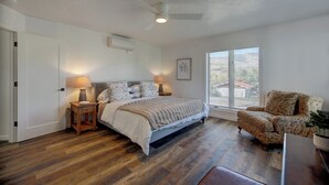 Ein Schlafzimmer mit Bett, Nachttischlampen, Deckenventilator, Fenster mit Jalousien und einem gemusterten Sessel.