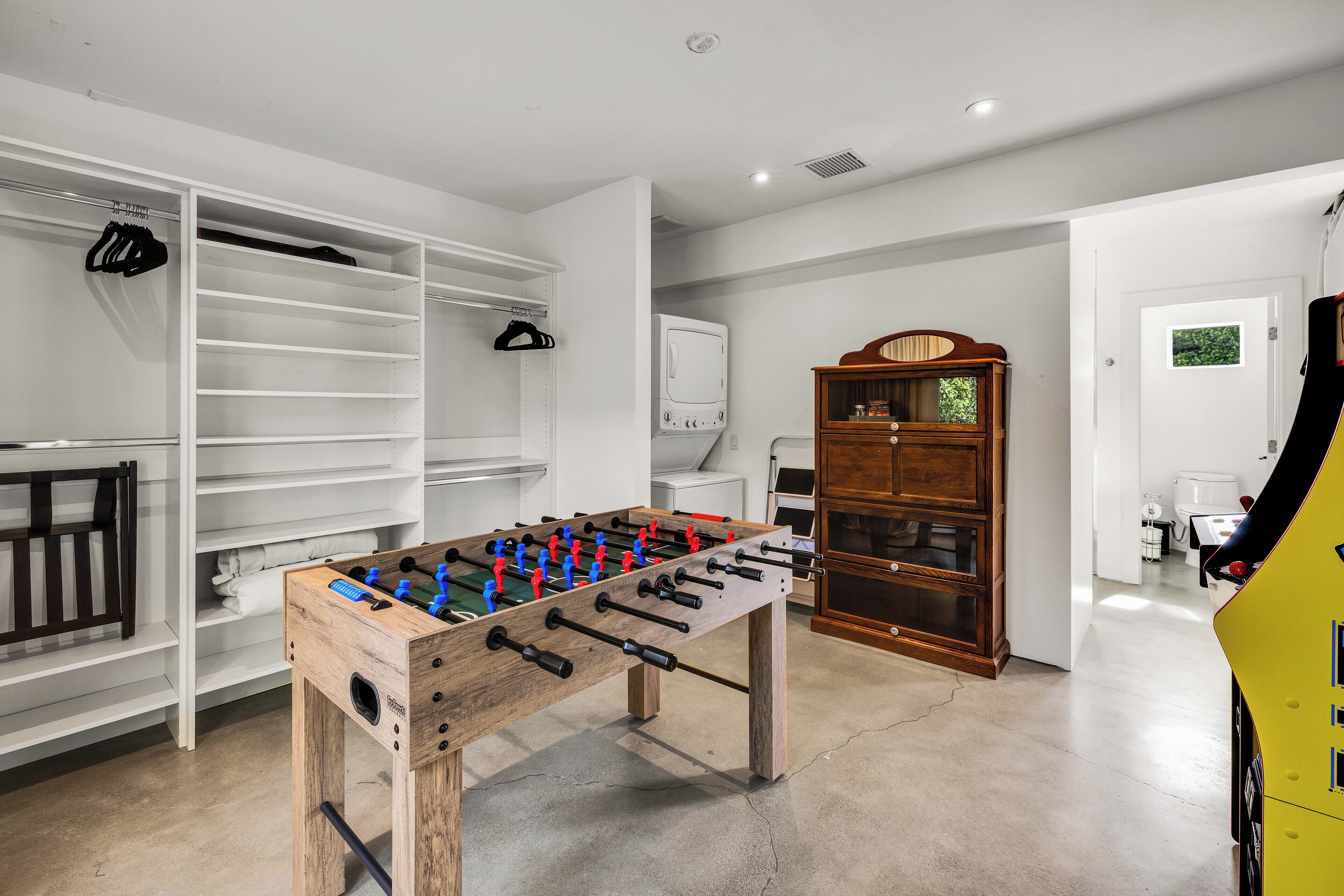 A room with a foosball table, a wooden cabinet, and a closet with shelves and hanging items.