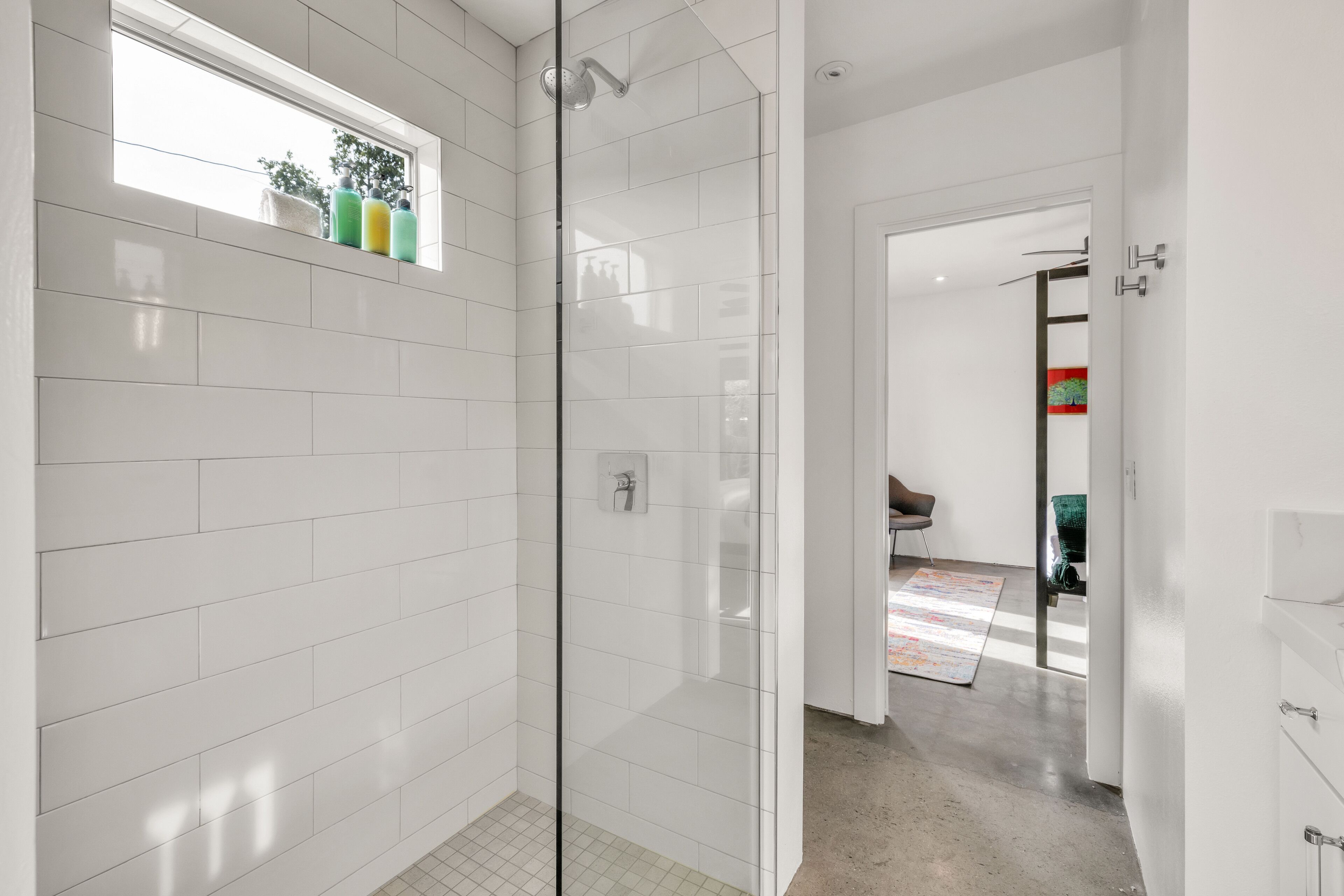 A modern bathroom with white tiles, a glass shower enclosure, and a window with potted plants.