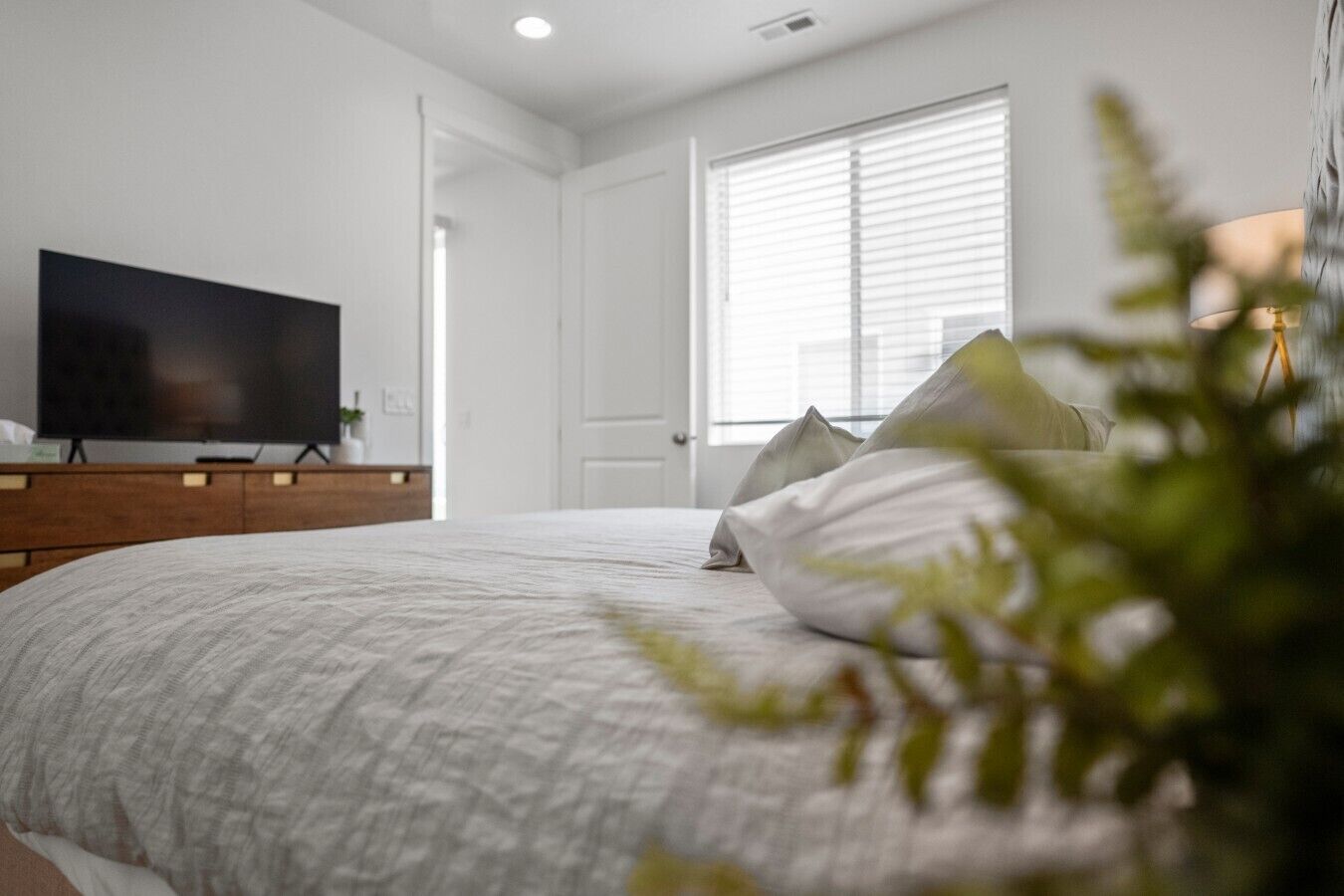 A bedroom with a bed, a television on a stand, a door, and a window with blinds.