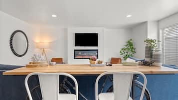 Une cuisine moderne avec un plan de travail en bois, deux chaises blanches, un téléviseur placé au-dessus du plan de travail et une plante sur la droite.