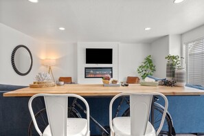 A modern kitchen with a wooden countertop, two white chairs, a television above the counter, and a plant on the right.