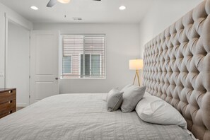 A bedroom with a tufted headboard, a bedside lamp, and a window with blinds. - Valley Views | Bear Lake Marina | King Suite + BBQ (Garden City)