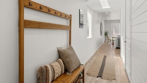 Un pasillo con un banco de madera, dos almohadas, una alfombra, un espejo y una puerta que da a otra habitación.