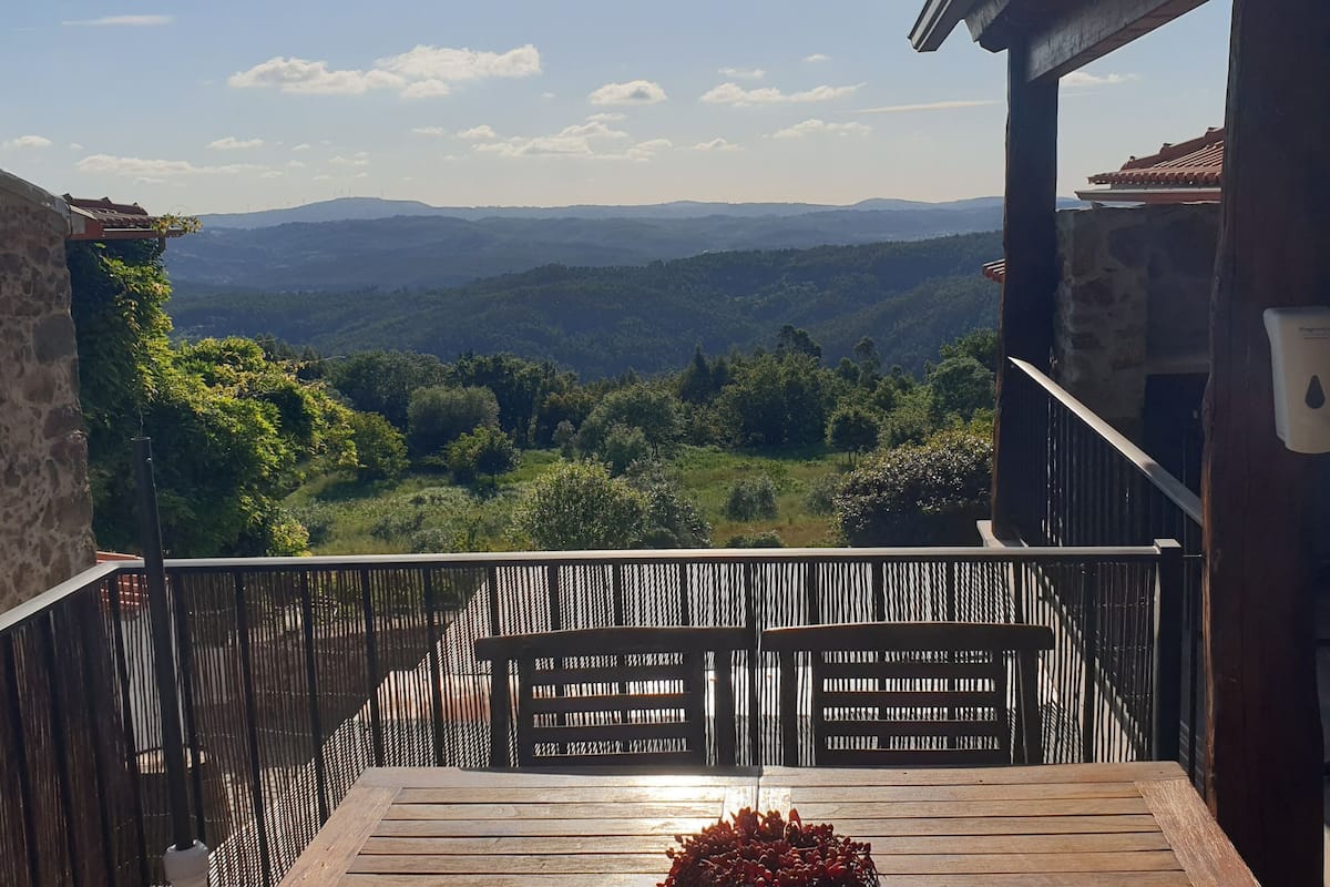 A balcony with a wooden table and chairs, a stone wall, and a view of a mountainous landscape.