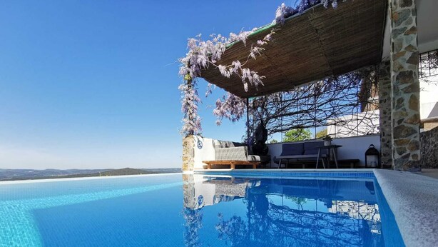 Una piscina infinita azul cristalina con vistas al mar, una pérgola adornada con flores blancas y un muro de piedra.