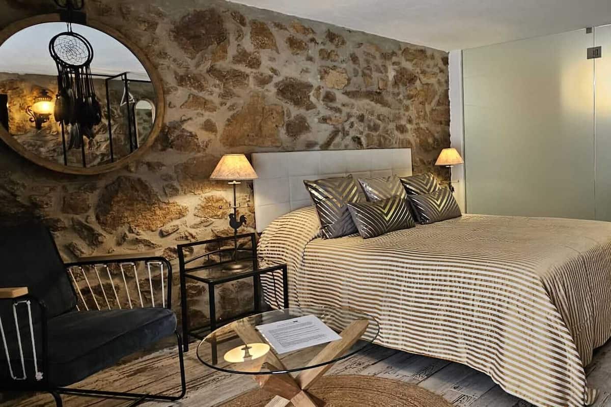 A bedroom with a stone wall, a bed with striped bedding, two bedside lamps, a round mirror, a small table with a lamp, and a bunk bed in the corner.