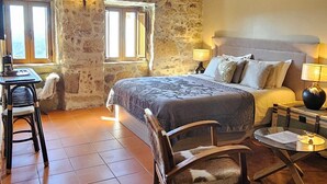 Un dormitorio rústico con vigas de madera a la vista, una pared de piedra, una cama con cabecero gris, una mesita con lámpara y una silla con una manta de piel.