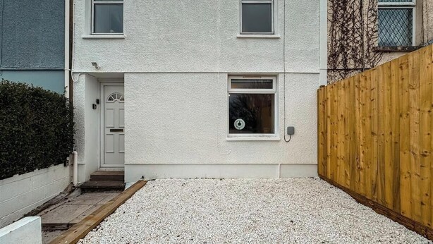 Un edificio residenziale a due piani con facciata bianca, una porta d'ingresso, due finestre, una recinzione in legno e un vialetto sterrato.