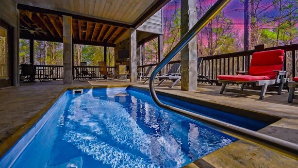 An indoor swimming pool with a wooden deck, lounge chairs, and a view of trees through large windows. - Merlot Mountain • Pool, Hottub, Firepit, Fireplace (Broken Bow)