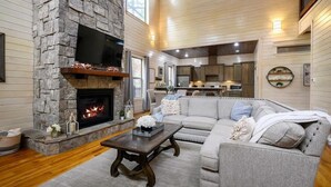 A cozy living room with a stone fireplace, a large TV, and a comfortable sofa. - Merlot Mountain • Pool, Hottub, Firepit, Fireplace (Broken Bow)