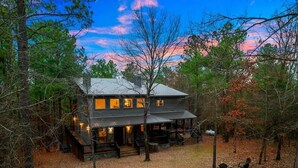 A two-story cabin with a porch, surrounded by trees and a leaf-covered ground. - Merlot Mountain • Pool, Hottub, Firepit, Fireplace (Broken Bow)