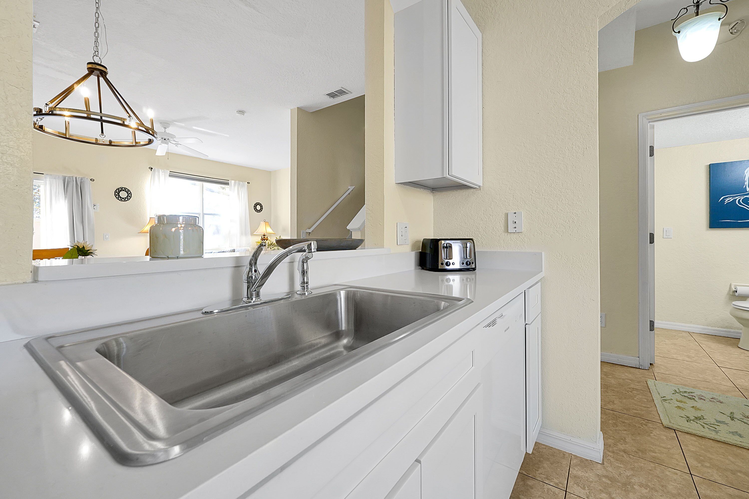 A kitchen with a stainless steel sink, white cabinets, a chandelier, and a view of the living area.