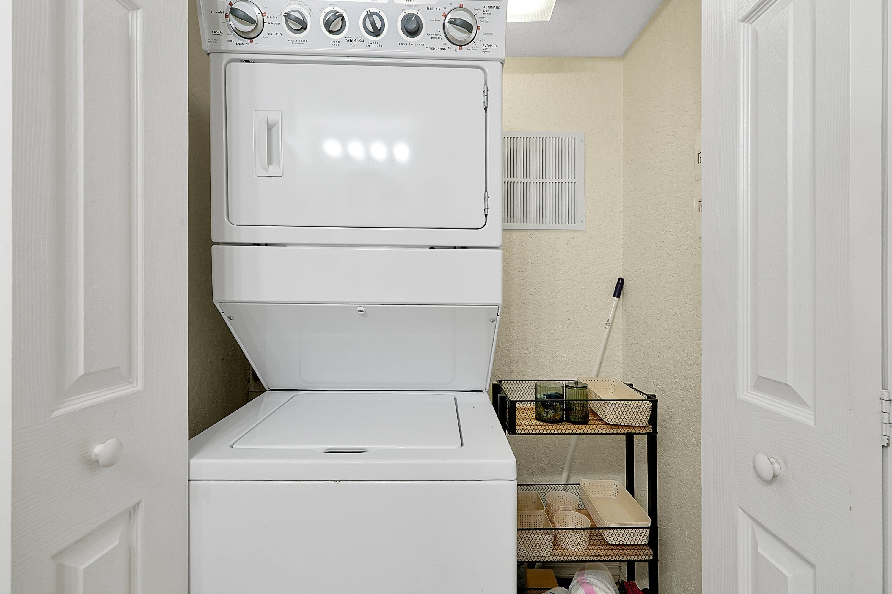 A stacked washer and dryer unit in a laundry room with a metal rack holding baskets and a power outlet on the wall.