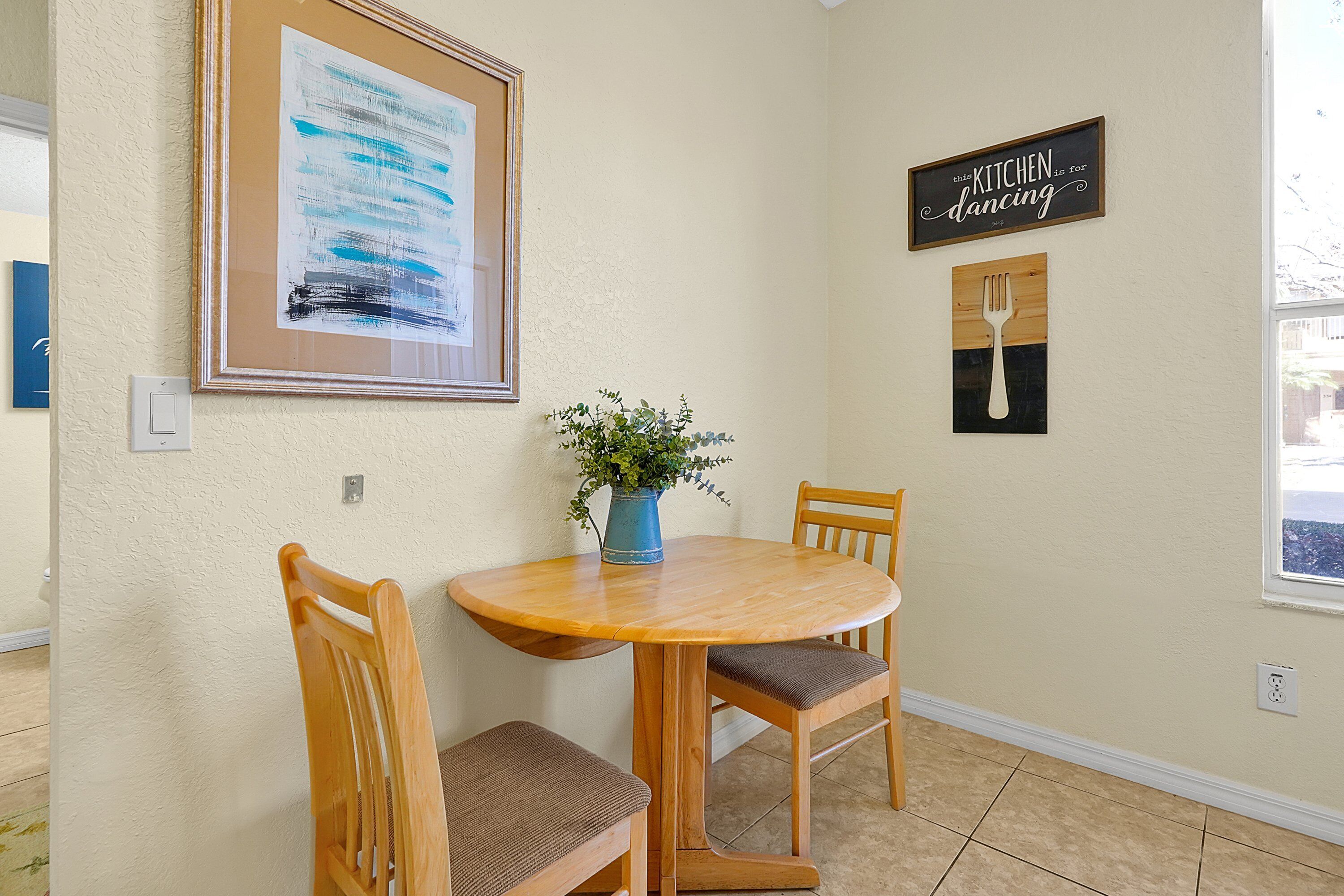 Un comedor con una mesa redonda de madera, dos sillas de madera, un florero con planta y adornos en la pared, incluyendo un cuadro abstracto enmarcado y un letrero que dice "Esta cocina es para bailar."