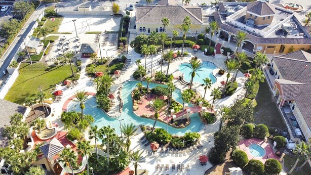 Eine Vogelperspektive zeigt einen luxuriösen Urlaubsort mit einem zentralen Pool, mehreren Liegebereichen und viel Grün umgeben.