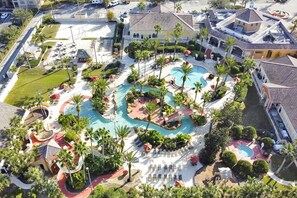 An aerial view of a resort with a central swimming pool, multiple lounge areas, and surrounding greenery.
