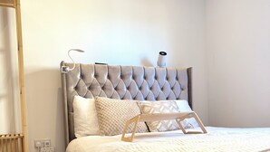 Une chambre à coucher bien aménagée, avec une tête de lit capitonnée, une table de chevet, un petit tabouret et une prise électrique au mur.
