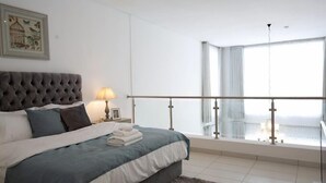 A modern bedroom with a tufted headboard, a large bed with a blue and white duvet, a nightstand with a lamp, and a balcony with a glass railing. - Shorlets by First Bridgeford Homes (Lekki)