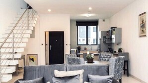 A modern living room with a grey sofa, a staircase, and a kitchen area in the background. - Shorlets by First Bridgeford Homes (Lekki)
