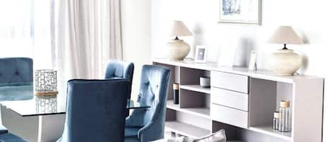 A modern living room with a glass coffee table, blue upholstered chairs, a grey sofa, and a white console table with lamps.