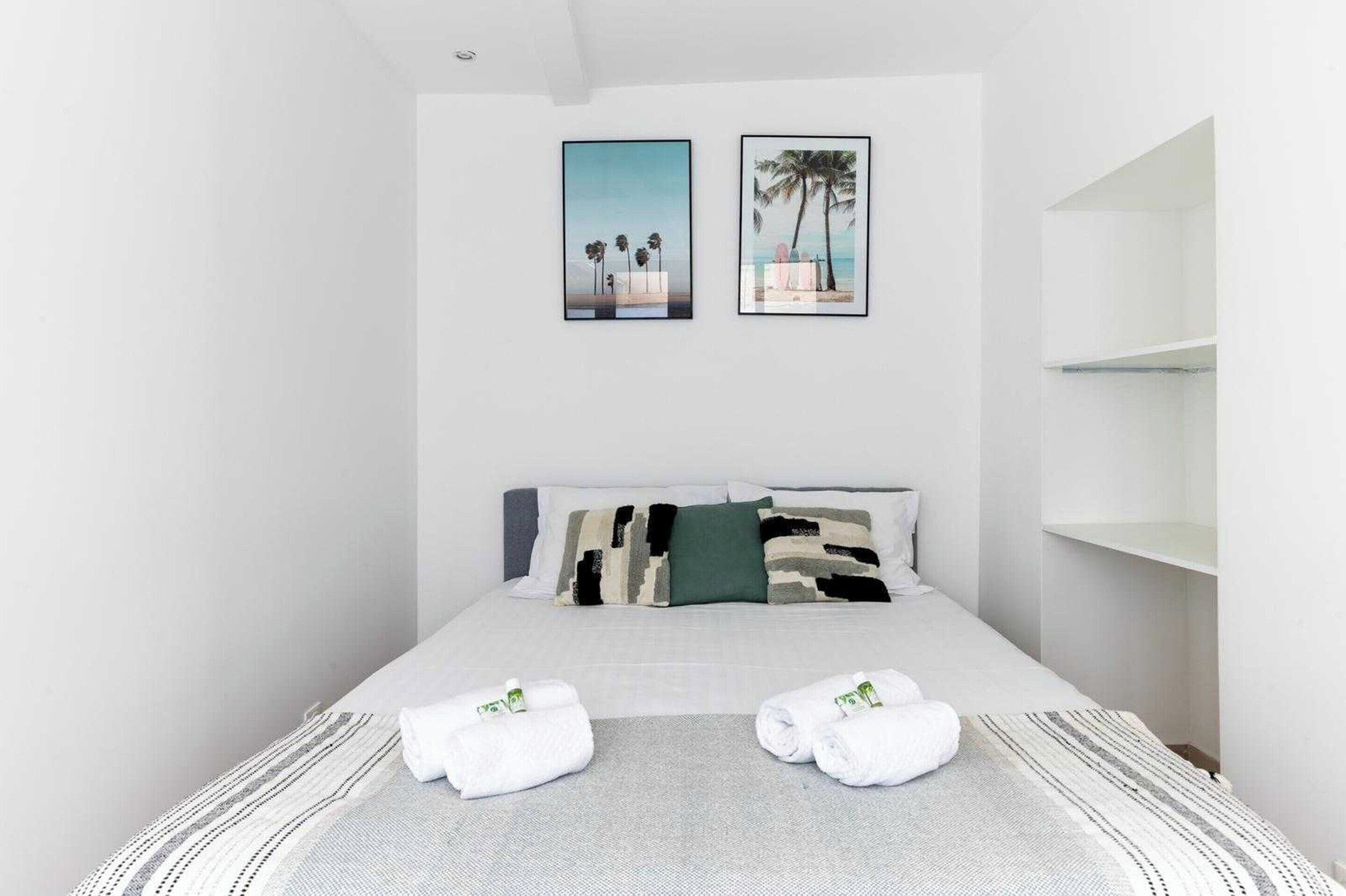 Habitación de hotel con cama, dos cuadros enmarcados, un estante blanco y dos toallas sobre la cama.