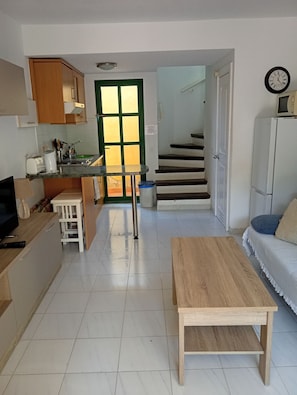 A compact living space with a kitchen area, a dining table, a sofa, and a staircase leading to an upper floor. - Duplex Apartment, with private garden, in quiet complex (Calete de Fuste)