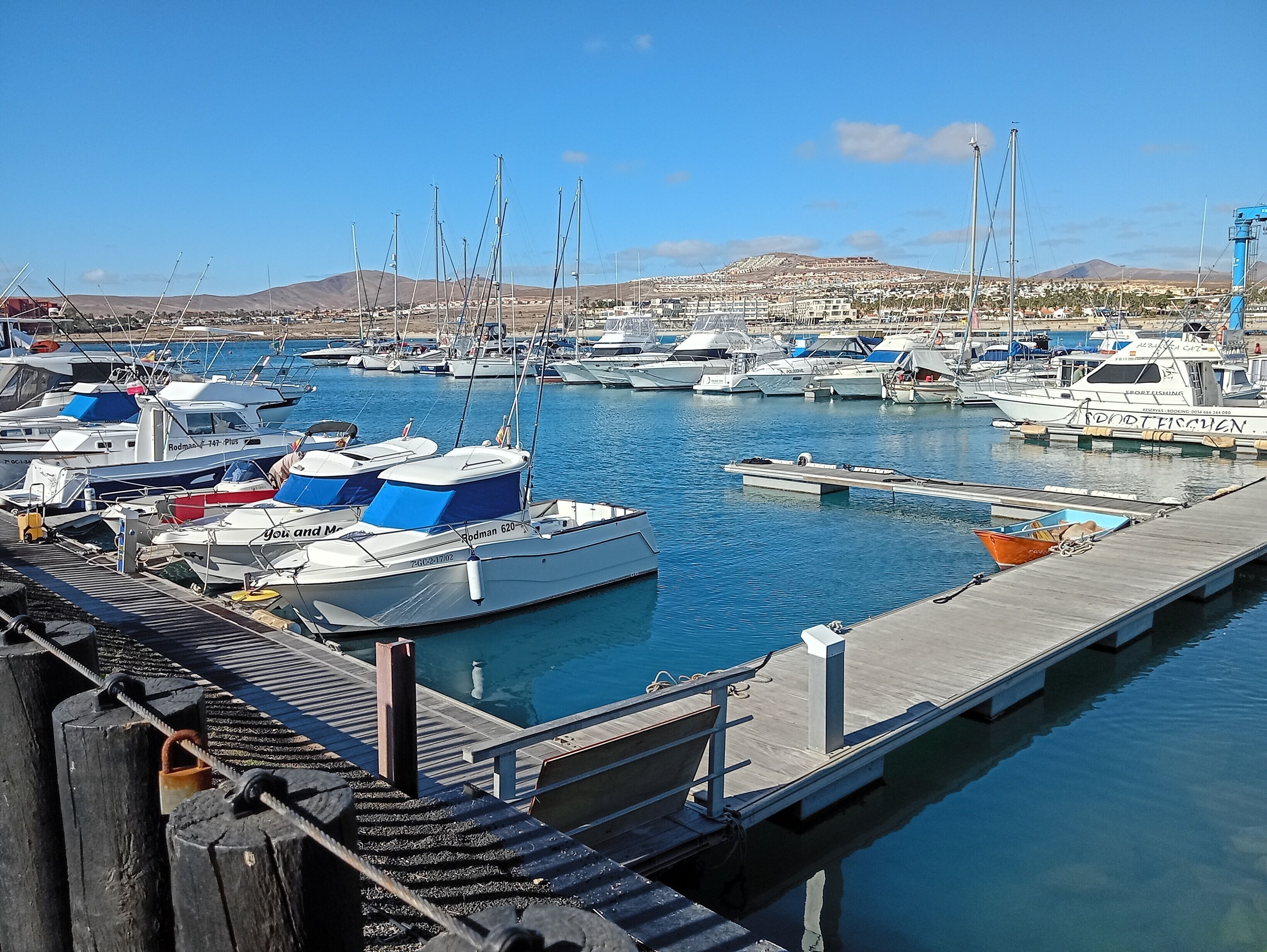 A marina with multiple boats docked, a wooden dock, and a mountainous landscape in the background.