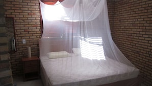 Un letto con copripiumino e cuscini bianchi, un comodino in legno, una parete in mattoni e una finestra con tenda leggera.