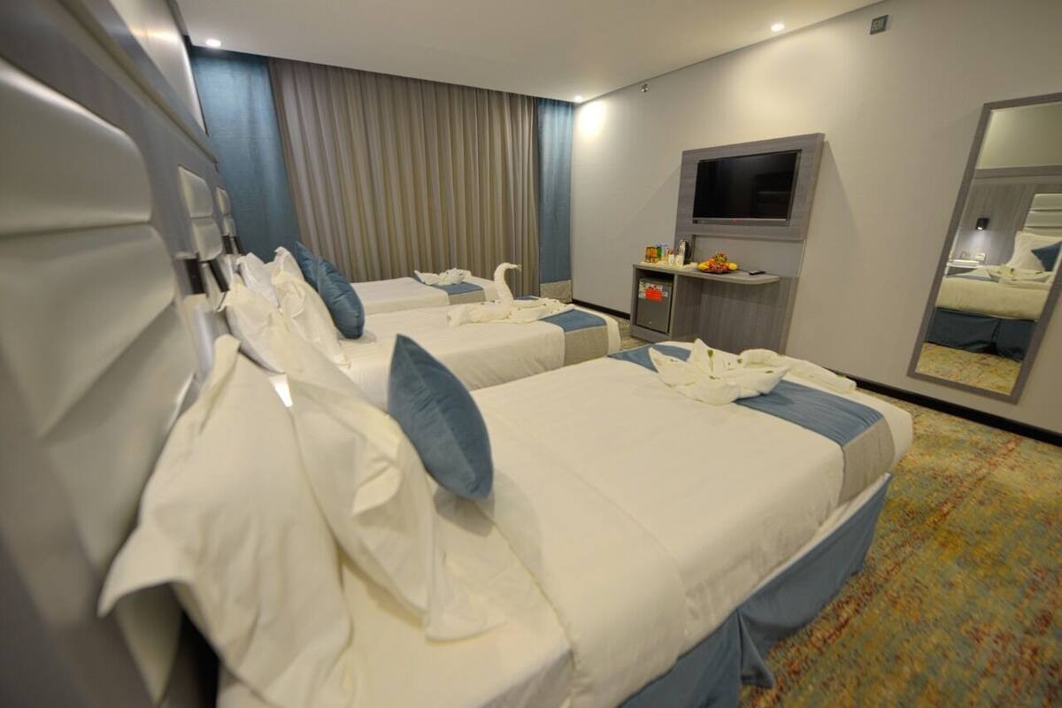 A hotel room with two beds, a TV, a small table with a fruit bowl, and a mirror.