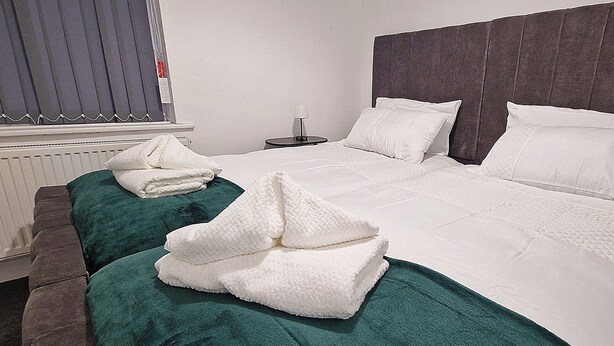Quarto de hotel com uma cama grande, duas almofadas, mesa de cabeceira com abajur e janela com persianas.
