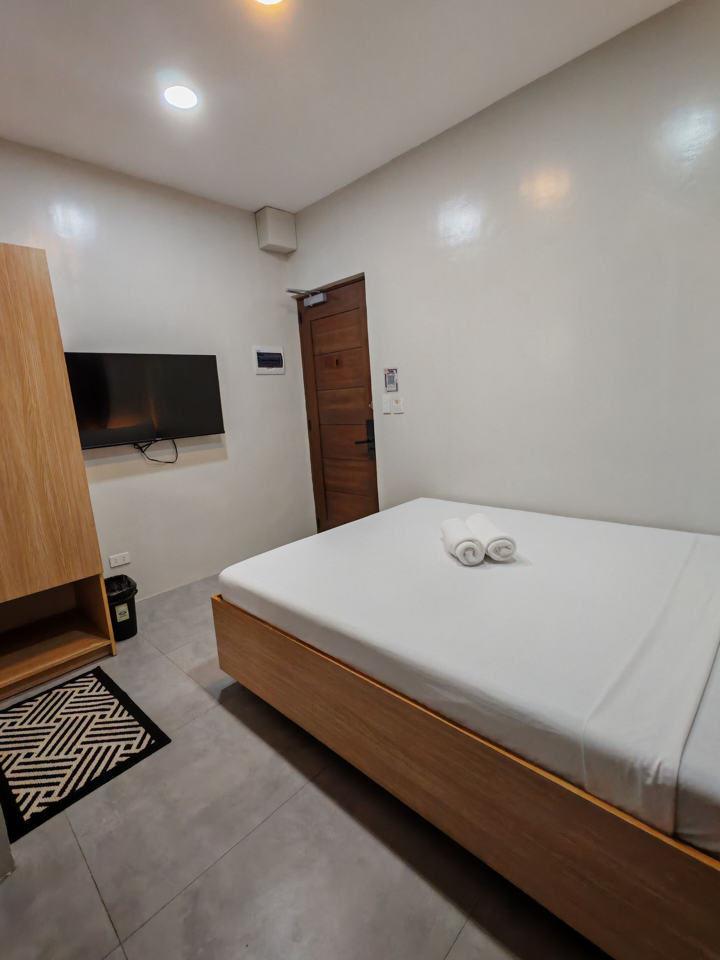 A modern hotel room with a wooden bed, a flat-screen TV, a wooden wardrobe, and a small window.