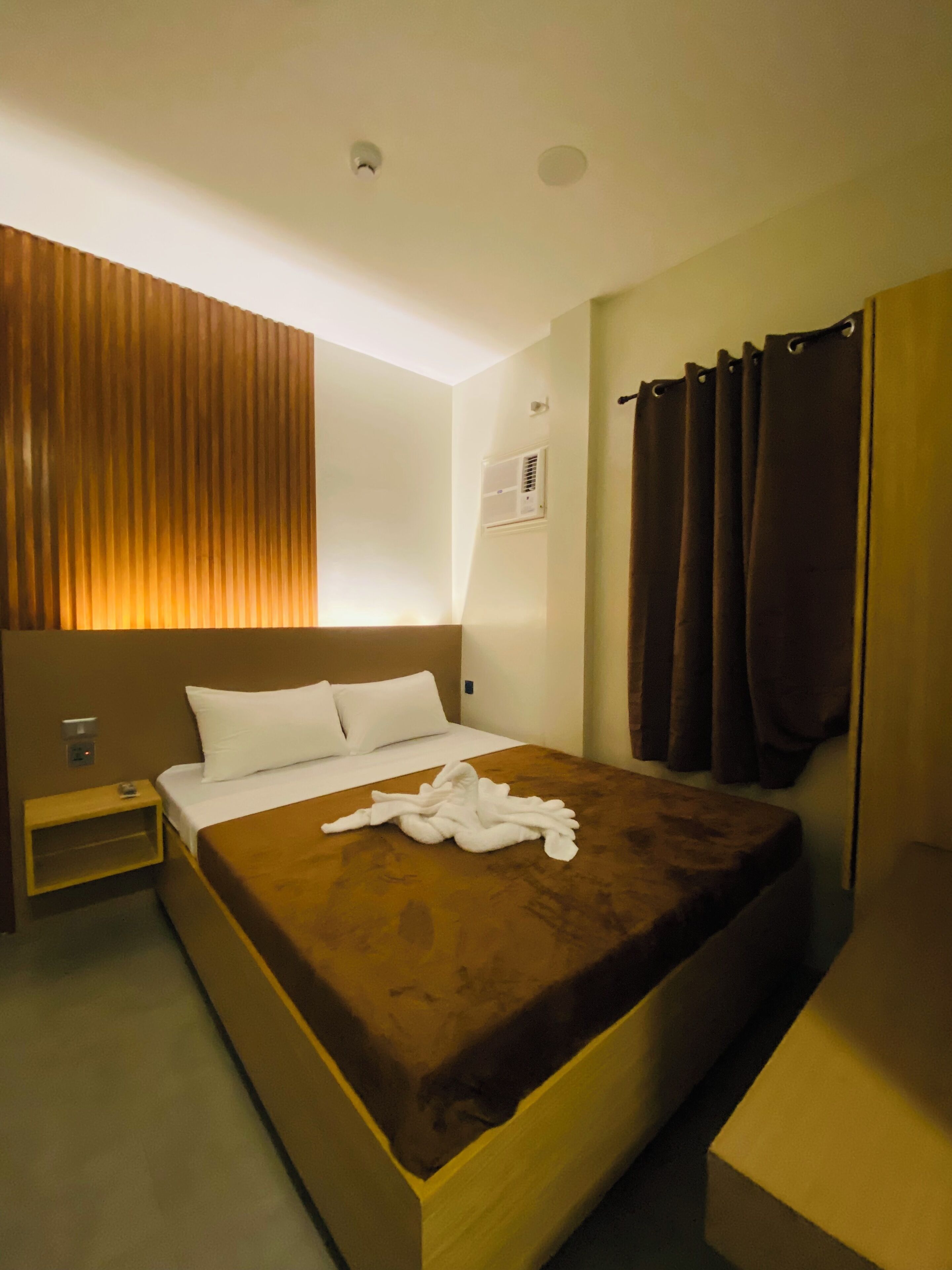 Habitación de hotel con una cama grande, una mesita de noche, aire acondicionado de pared y una ventana con cortinas.