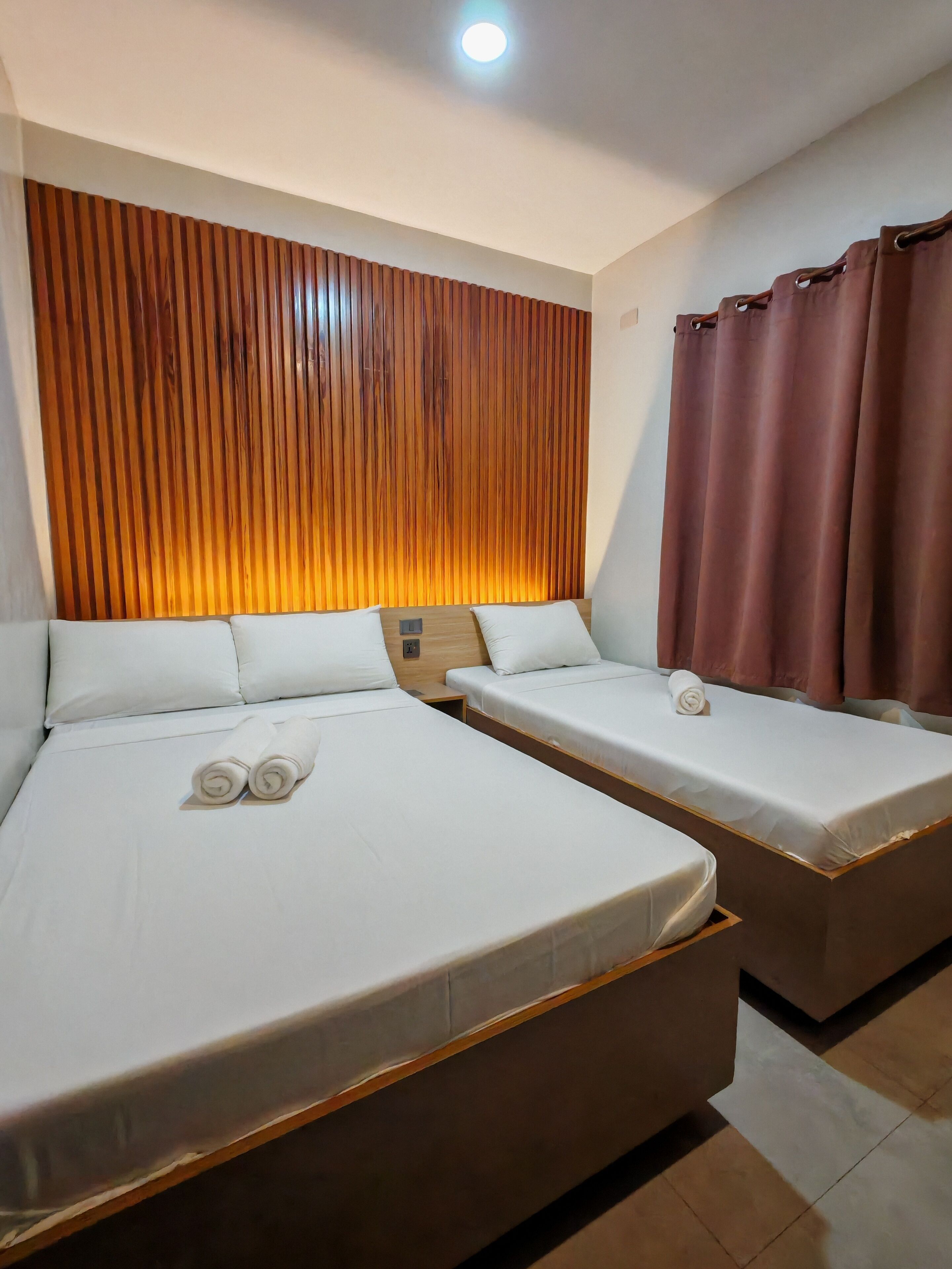 Habitación de hotel con dos camas, cabecero de madera, cortina marrón y una lámpara en el techo.