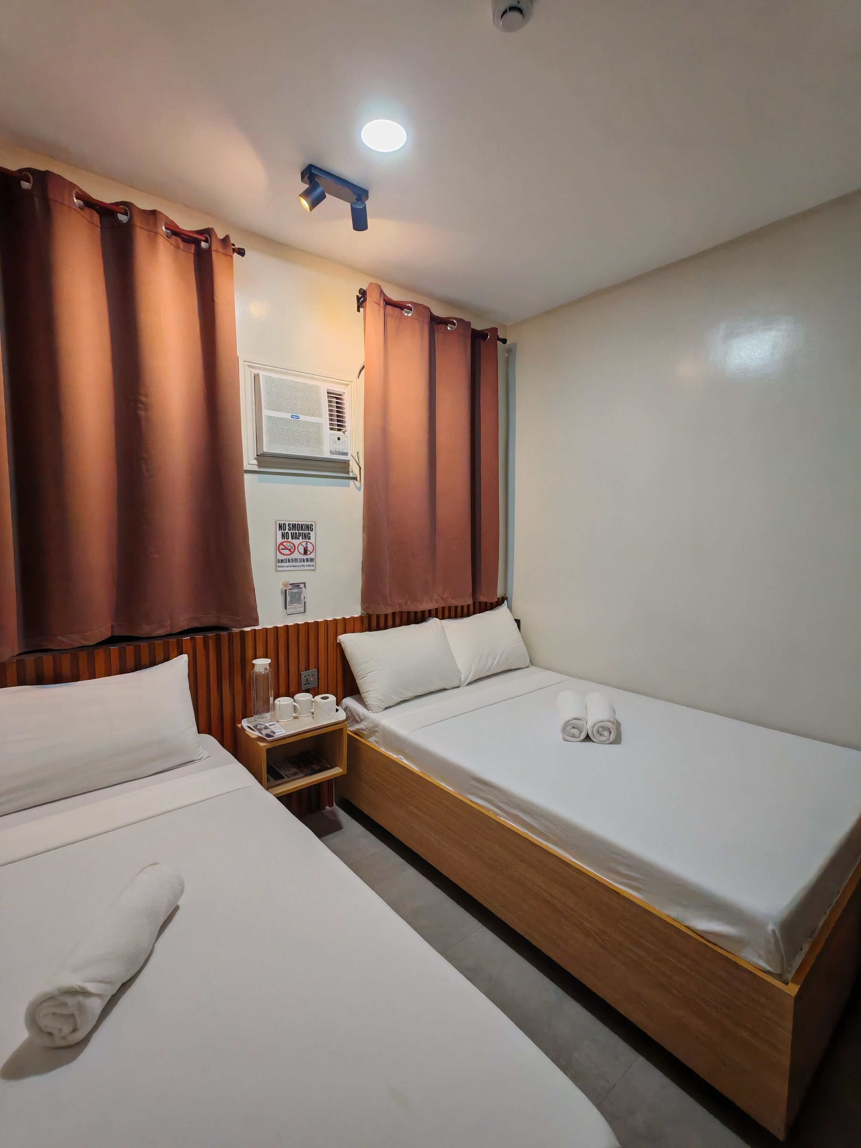 A hotel room with two beds, a nightstand, a wall-mounted air conditioning unit, and a no-smoking sign.