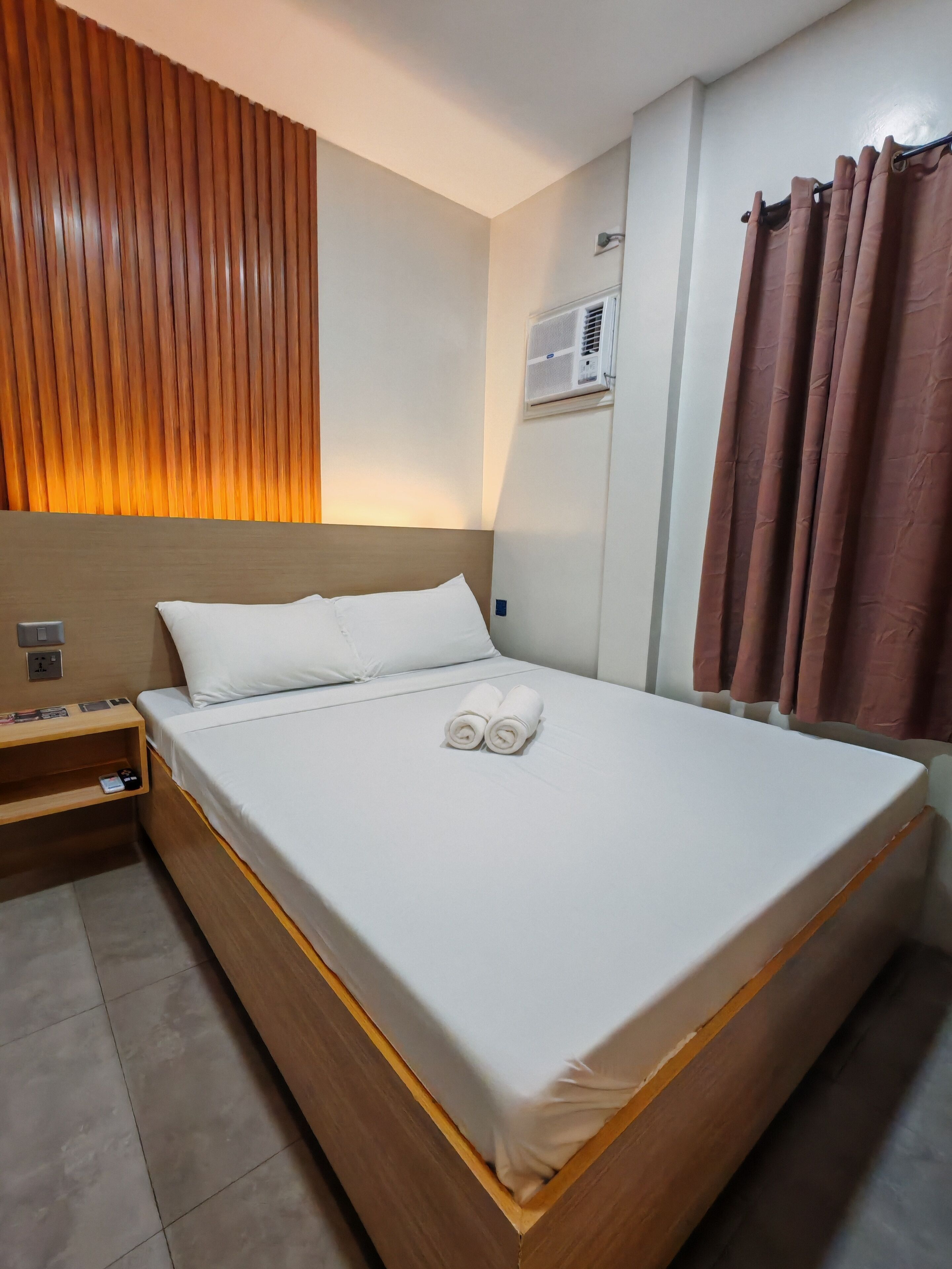 A neatly made bed with white linens and two rolled towels, a wooden headboard, a nightstand with a lamp, and a wall-mounted air conditioner.
