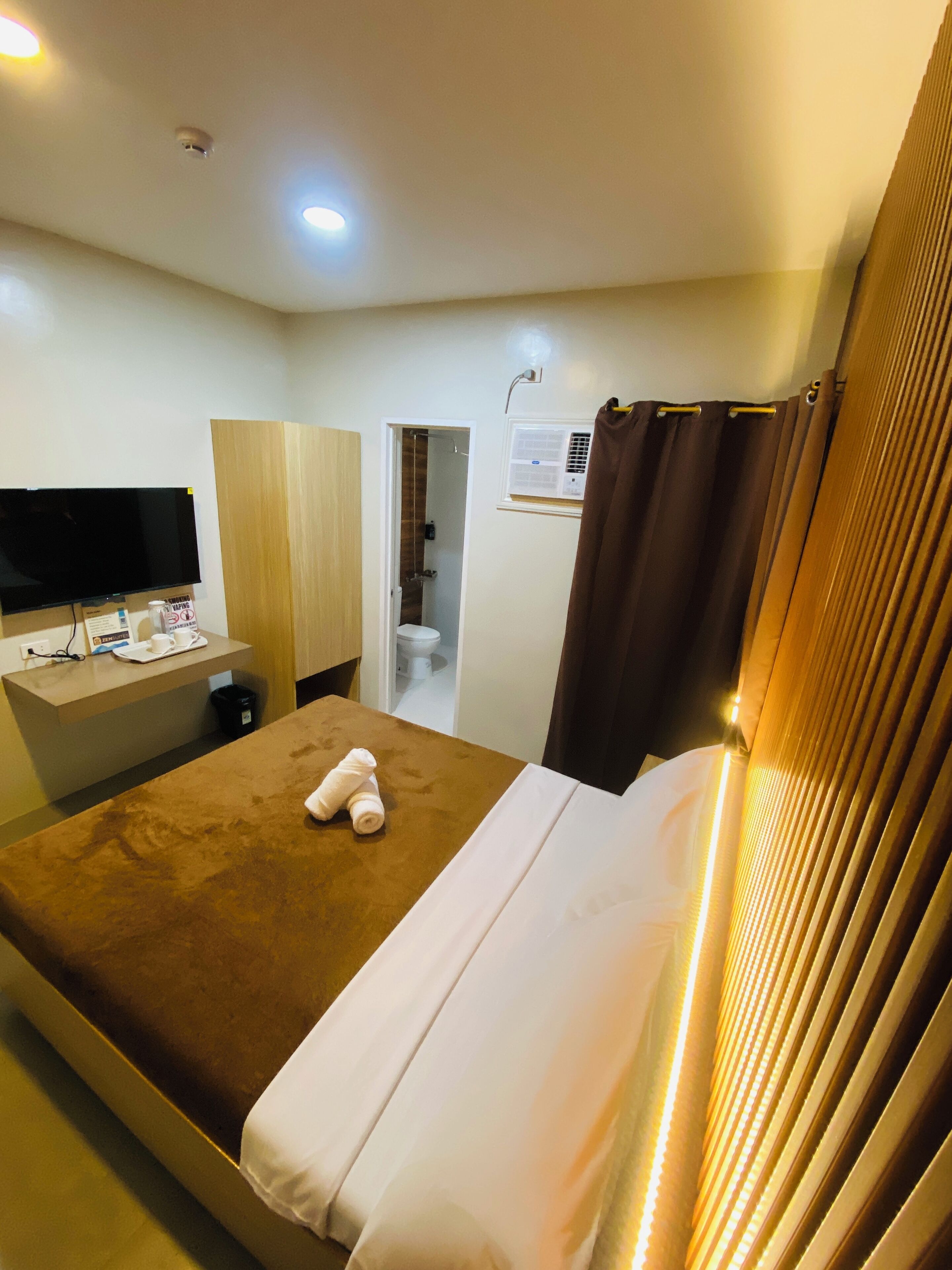 A hotel room with a bed, a desk with a flat-screen TV, a bathroom with a toilet and shower, and a window with curtains.