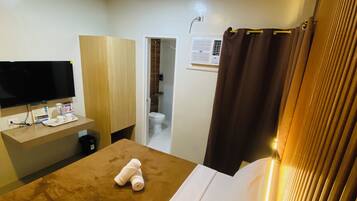 A hotel room with a bed, a desk with a flat-screen TV, a bathroom with a toilet and shower, and a window with curtains.