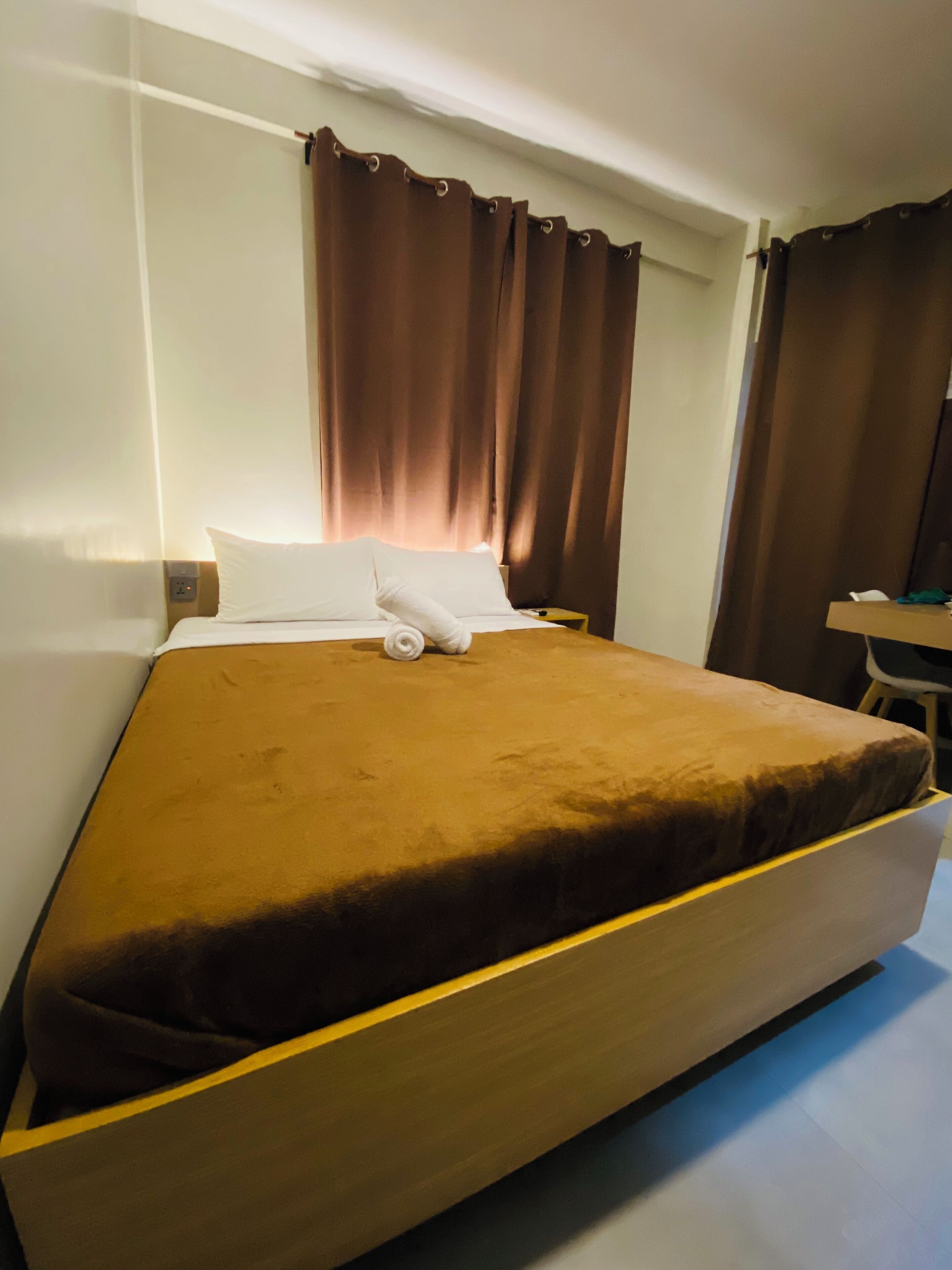 A hotel room with a large bed, brown curtains, a wooden headboard, and a small bedside table.