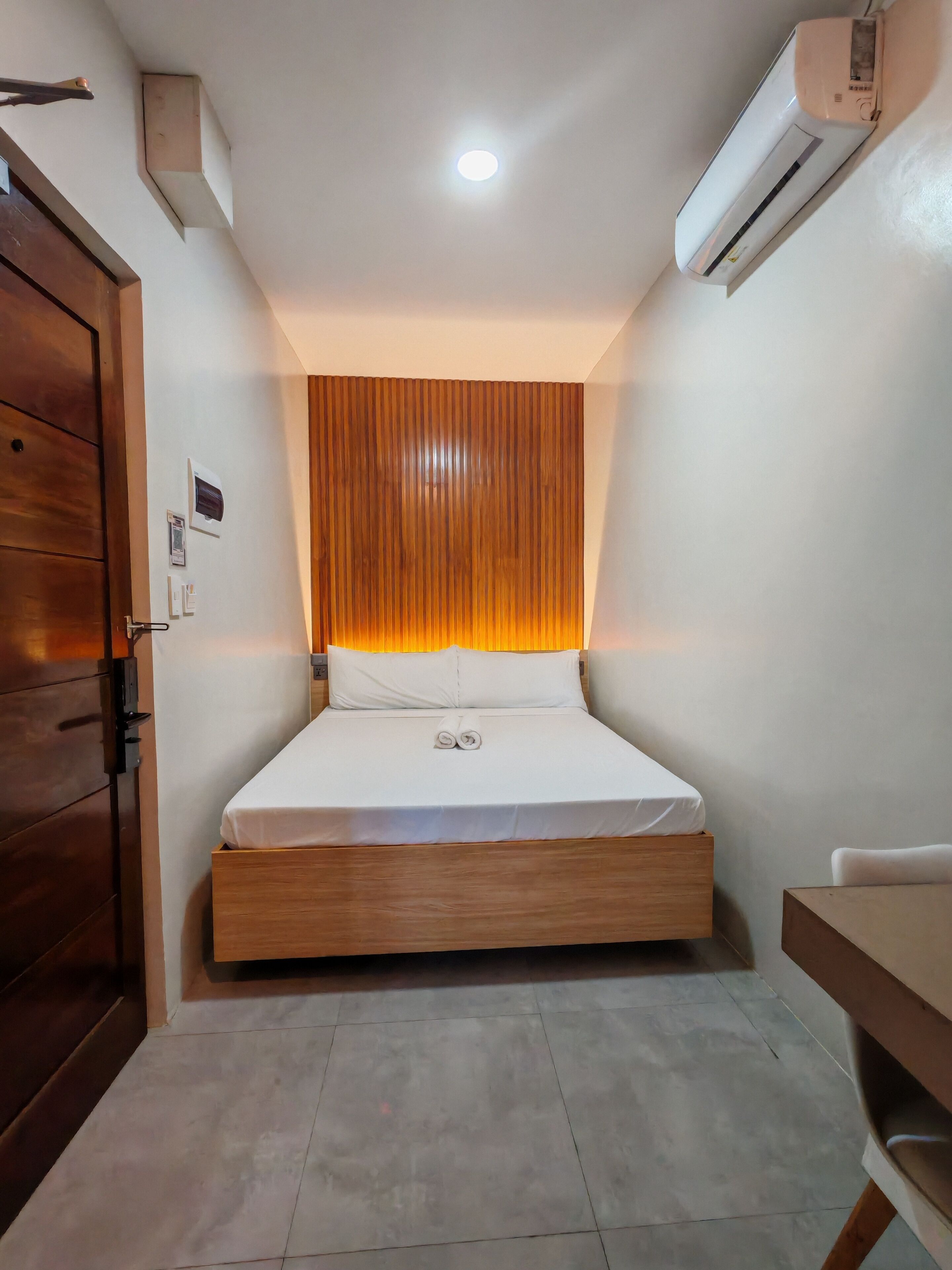 A hotel room with a wooden bed frame, a white mattress, a wooden headboard, a wooden door, and an air conditioning unit on the wall.