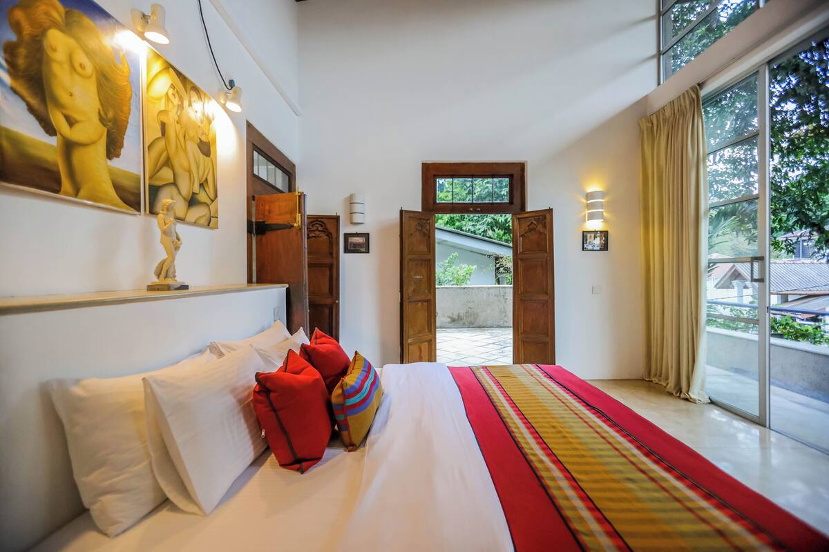 A hotel room with a large bed, a view of the outside, and artwork on the wall.