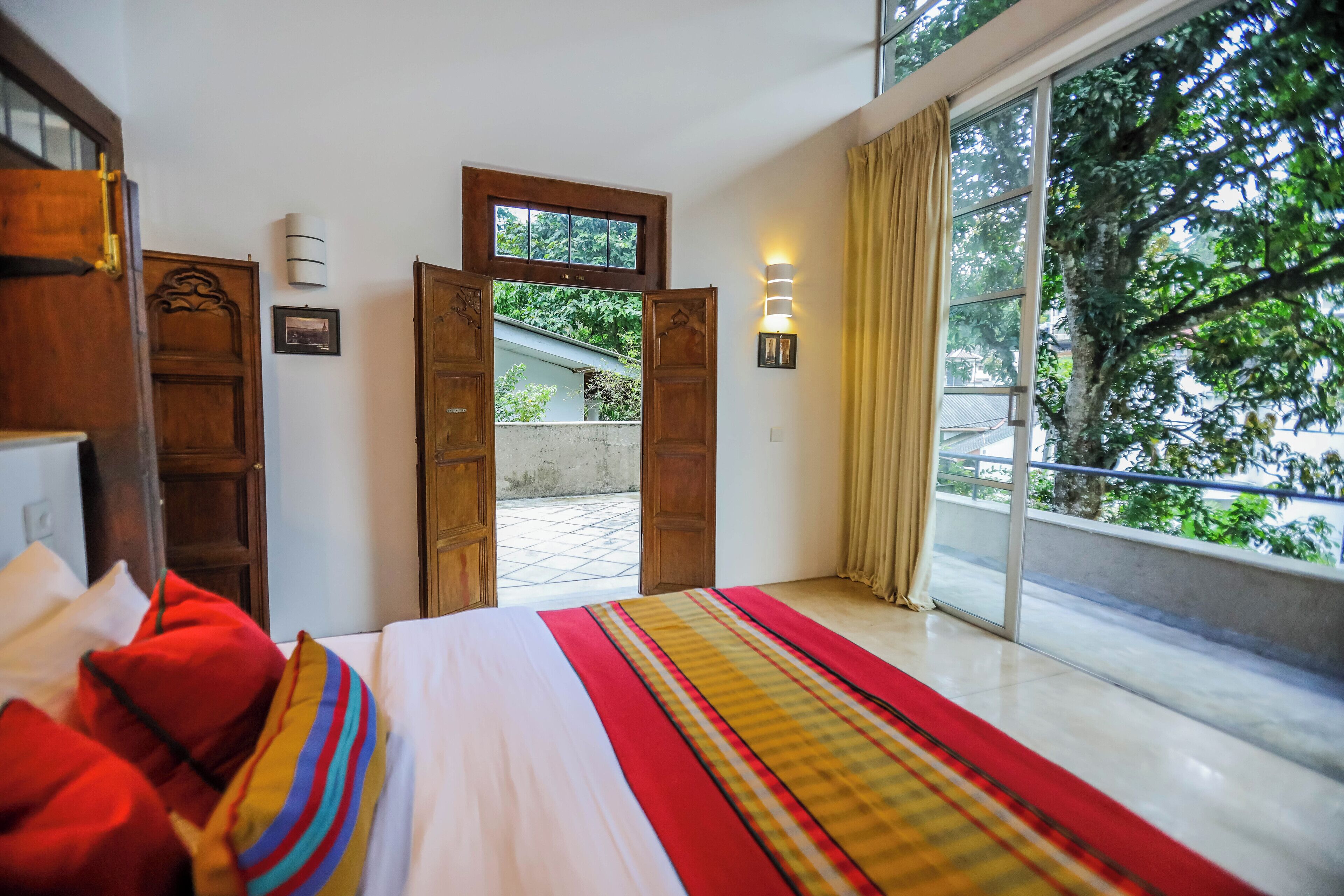 A bedroom with a large bed, wooden doors, a window with curtains, and a view of greenery outside.