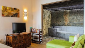 A living room with a television, bookshelf, green sofa, and a stone wall with a bathtub. - Margossa Residence Kandy - Two Bedroom Design Villa (Kandy)