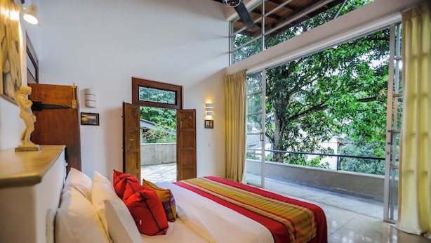 A hotel room with a large bed, a wooden headboard, a window with a view of trees, and a balcony. - Margossa Residence Kandy - Two Bedroom Design Villa (Kandy)