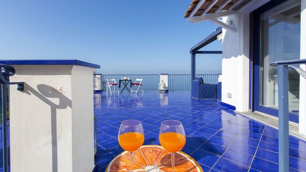 A balcony with a blue tiled floor, two glasses of orange juice on a table, and a view of the sea. - Villa Blue Capri in Massa Lubrense (Massa Lubrense)